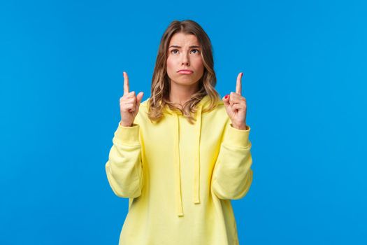 Jealous and gloomy cute blond female student feel regret not having something she desires, sobbing frowning sad as looking pointing up, standing over blue background.