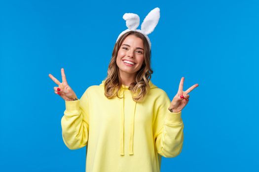 Holidays, traditions and celebration concept. Kawaii young blond girl in rabbit ears showing peace gesture and smiling, having fun, enjoying party, standing blue background.