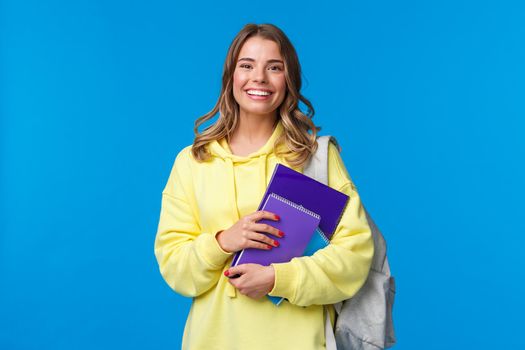 Cheerful pretty blond girl smiling at camera, carry backpack and notebooks, papers for studying, learning new language at courses, standing joyful over blue background. Education and people concept