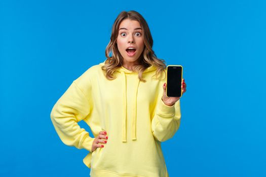 Impressed and surprised cute european girl showing amazing new app or mobile game, holding smartphone facing display to camera, staring speechless with excitement, blue background.