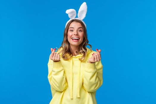 Holidays, traditions and celebration concept. Cheerful european girl celebrating Easter day, wearing rabbit ears show korean hearts sign and smiling joyfully, stand blue background.
