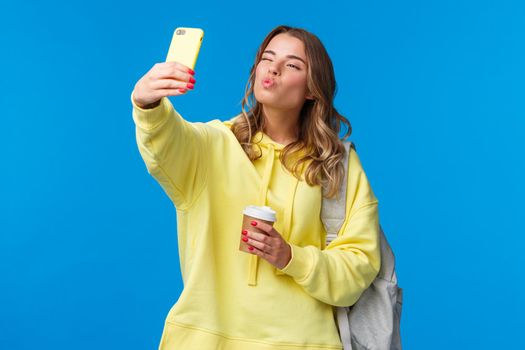 Cute positive and feminine beautiful young female student heading college holding paper cup from favorite cafe and backpack with books, taking selfie show kiss mwah face at mobile camera.