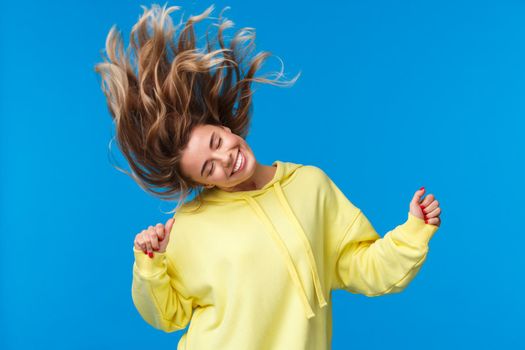 Carefree relaxed and joyful beautiful blond girl shaking head, dancing and jumping from happiness, having fun at awesome party or music concert, standing blue background.