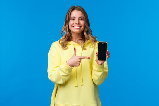 Proud cheerful young blond woman advertise new mobile game or application, look camera and smiling white teeth as pointing smartphone display, recommend download or try out.