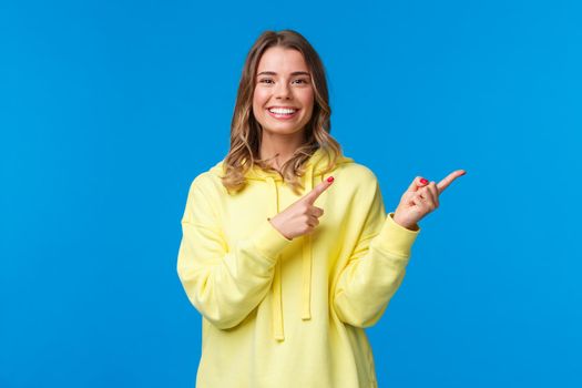 Good-looking friendly pleasant blond girl in yellow hoodie pointing fingers right and smiling white teeth at camera as recommend place, asking visit or click link, standing blue background.