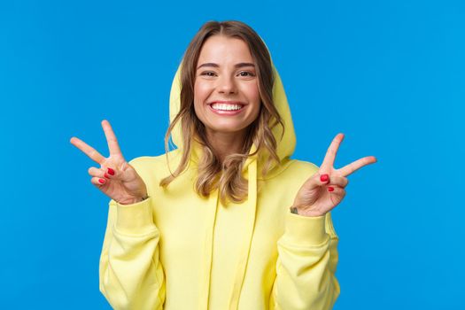 Close-up portrait cute silly blond european female in yellow hoodie, showing peace gesture and smiling optimistic, send positive vibe, standing blue background upbeat, make kawaii pose.