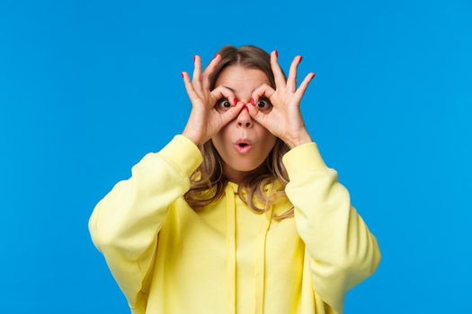Impressed and intrigued, curious young caucasian girl in yellow hoodie looking at something awesome, found great product special discount, make glasses with fingers on eyes, blue background.