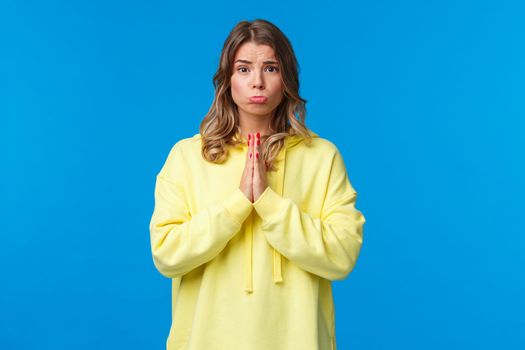 Cute silly clingy girlfriend asking to buy her something she wants while discount on, hold hands in pray pouting and looking camera pleading, begging for favour, stand blue background.