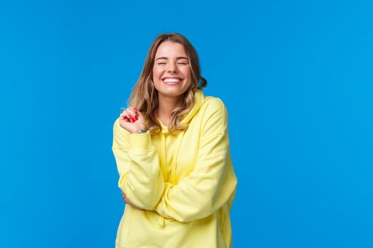 Cheerful carefree pleased girl feel happy and delighted, close eyes dreamy smiling with perfect beaming grin, touch strand of hair, standing in yellow hoodie over blue background.