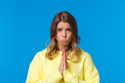 Close-up portrait of cute sulky european female blonde begging for help, asking favour, really need something, holding hands in pray, frowing and sobbing as plead for apology, blue background.