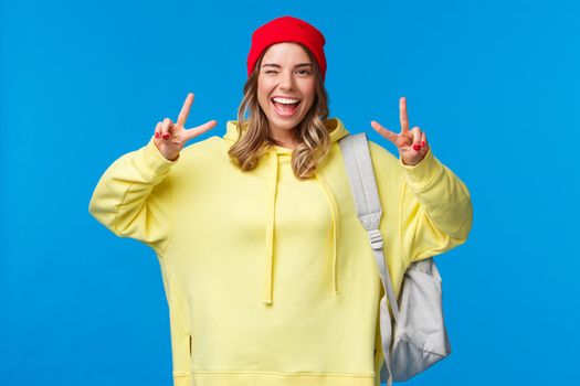 Back to school concept. Cheerful hipster girl with blond hair in beanie, carry backpack, wink carefree and smiling, showing peace gesture send positive vibes, stand blue background.