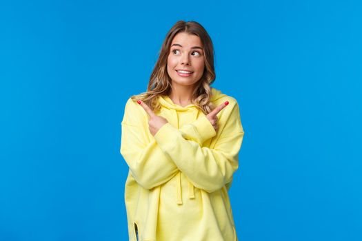 Various choices, cant decide. Cute dreamy blond girl in yellow hoodie making her choice, pointing fingers sideways at left and right side and smiling, picking one thing from all, blue background.