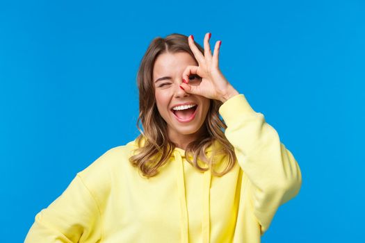 Close-up portrait of optimistic dreamy caucasian girl with blonde short hair, showing okay gesture over eye and wink happy, express positive enthusiastic emotion, standing blue background.