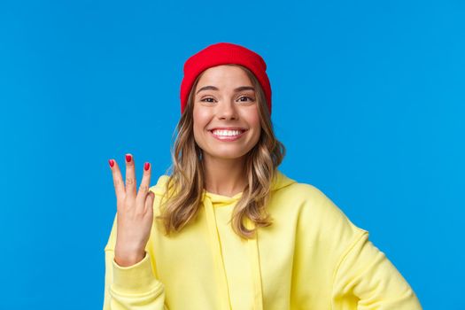Good-looking caucasian female in red beanie and yellow hoodie, showing number three and smiling, make order or reservetion, standing proud and delighted, blue background. Copy space