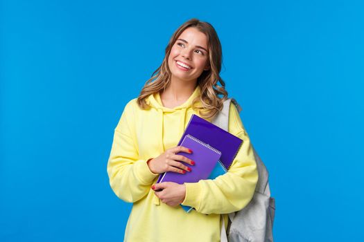 Dreamy happy good-looking pretty blond girl studying in college, holding backpack and notebooks, homework staff, look upper left corner thoughtful, standing blue background.