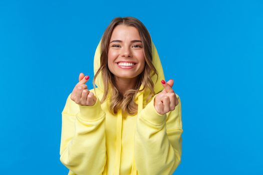 Close-up portrait of cute and silly blond caucasian girl in yellow hoodie, showing korean heart gesture and smiling, vibing, enjoying party, having fun over blue background. Copy space