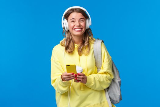 Cheerful pretty young female student with backpack, listening favorite playlist in headphones, laughing and smiling at camera carefree, hold mobile phone, stand blue background.