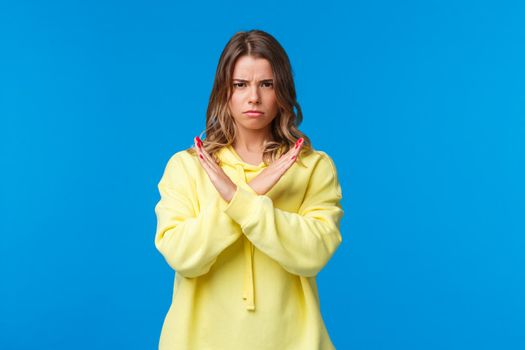 Stop right there, no means no. Serious-looking confident blond woman in yellow hoodie making cross on chest, enough and decline gesture, demand quit it, stand blue background.