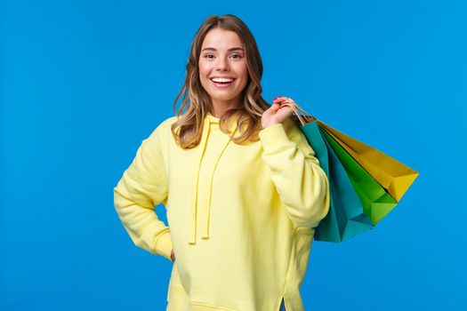Shopping time. Girl relaxing after hard working days buying things in store on weekend, hold bags on back and smiling relieved, release stress through purchasing clothes, blue background.