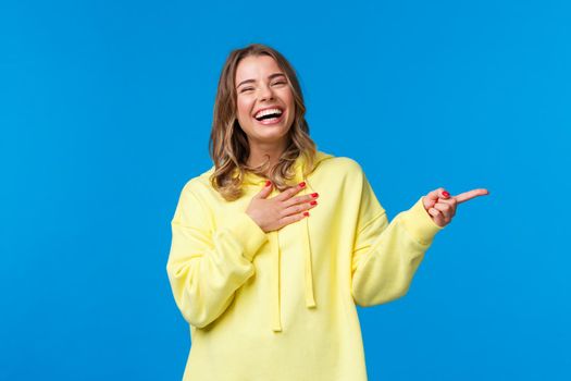 Waist-up portrait of laughing girl having fun, discuss hilarious stories with friends, chuckle touch chest as giggle carefree, look camera and smile as pointing finger right, blue background.