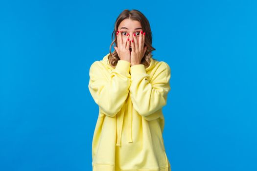 Shocked and speechless cute shy female cover eyes with hands as see naked guy scream and peeking through fingers with surprised startled expression, standing blue background.