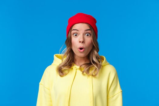 Wow tell me more. Intrigued and curious blond european girl staring astonished, gasping impressed, watching something exciting, hear interesting news, standing in red beanie and yellow hoodie.