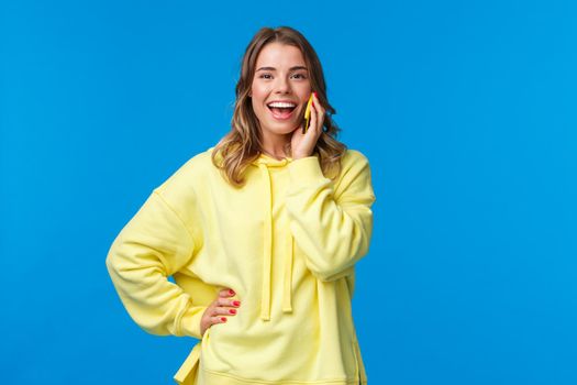 Carefree good-looking european blond girl in yellow hoodie lively talking on mobile phone, smiling camera as booking table for friends in bar, order food delivery, blue background.