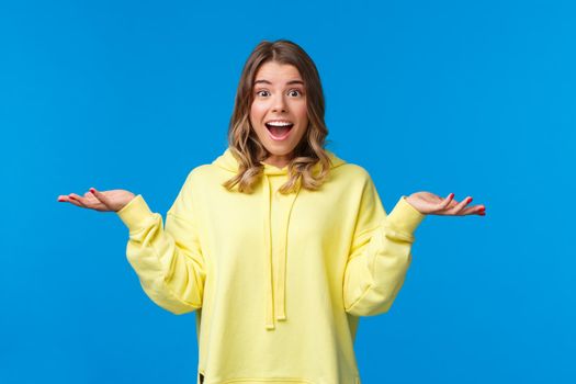 What nice surprise. Amused and happy cute blond caucasian woman spread hands sideways and shrugging smiling with excitement and disbelief, hear unexpected good news, blue background.
