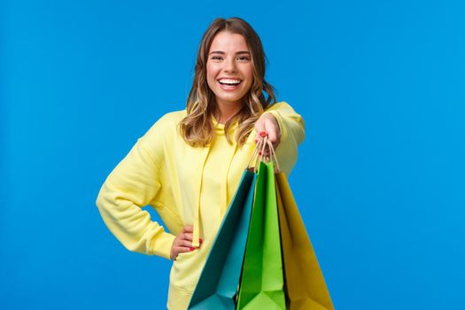 Cheerful blond girl giving you shopping bags and smiling happy, buying presents for family holiday, buying lots of staff in store and asking carry her packages, stand blue background.