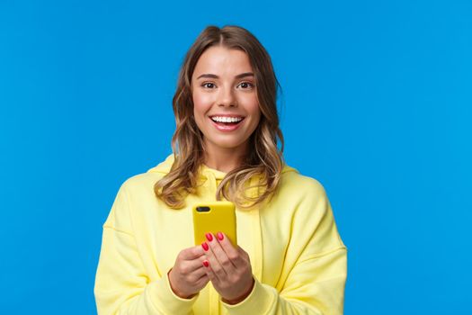 Close-up portrait excited smiling cute european female with blond haircut, holding mobile phone and looking camera entertained, standing blue background messaging, talking with friends online.