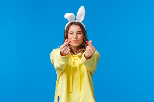 Holidays, traditions and celebration concept. Silly and cute happy blond girl having fun, showing korean heart gesture with fingers and send kisses, standing in rabbit ears, yellow hoodie.