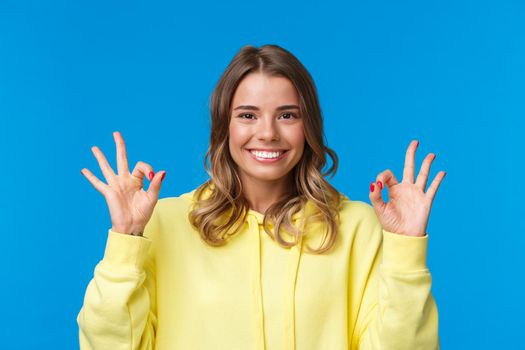 Close-up portrait happy confident young blond girl assure all okay, guarantee plan went well, smiling and show ok gesture in approval, confirmation or like, stand blue background. Copy space