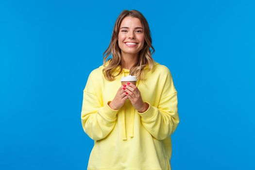 Girl enjoying tasty cup of morning coffee from favorite cafe. Cheerful lovely blond woman in yellow hoodie smiling white teeth and holding paper cup of take-away tea, stand blue background.