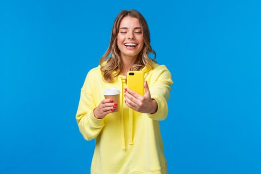 Cheerful smiling blond caucasian female blogger record video or take selfie on her smartphone, laughing and grinning as holding take-away cup of coffee, stand blue background.