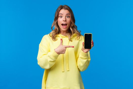 Impressed happy cheerful blond girl explain something as pointing mobile phone display, talking about new application or sharing her vacation photos, hold smartphone, look camera.