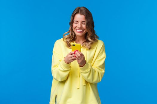 Joyful caucasian female with beautiful smile in yellow hoodie, holding mobile phone and laughing as texting friend hilarious story, send funny video, messaging or have conversation.