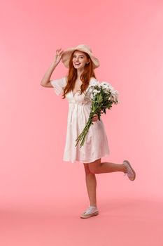 Full-length vertical shot excited and happy, cheerful redhead girl jumping from happiness and joy, overwhelmed with love, holding bouquet, smiling camera, received beautiful flowers, pink background.
