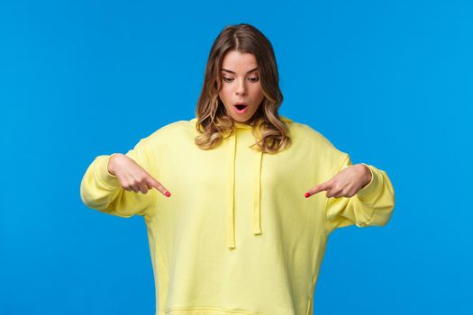 Girl found something stunning. Amused and speechless surprised blond alluring woman in yellow hoodie, drop jaw stare and pointing fingers down at cool product, blue background.