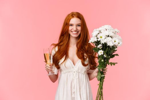 Attractive and excited romantic redhead girlfriend receive gift, holding white bouquet flowers and glass champagne, drinking celebrating occasion, having perfect valentines date, pink background.