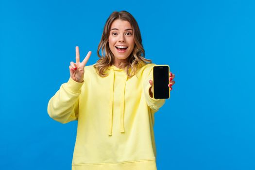 Optimistic cute blond european girl with short haircut, yellow hoodie, showing kawaii peace gesture and mobile phone display as using photo filter to edit and post pic online, blue background.