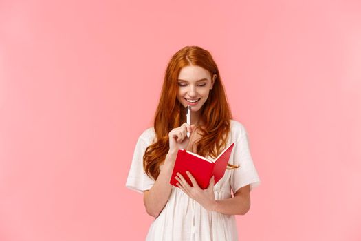 Thoughtful and happy, smiling pretty redhead female in white dress, touching chin with pen while thinking, making grocery or to-do list, writing something in cute red notebook, pink background.