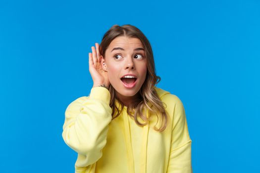 Close-up portrait of intrigued and curious blond young girl with pierced ear, gossiping, eavesdropping and listening to someone conversation interested, standing blue background. Copy space