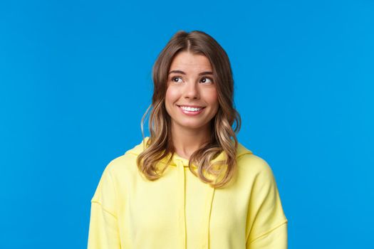 Close-up portrait of attractive european blond female with short hair, wear yellow hoodie, looking dreamy and silly upper left corner, smiling with white beaming smile, standing blue background.