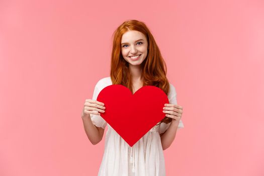 Tenderness, love and romance concept. Romantic charming redhead girlfriend prepared cute gift valentines day, showing red heart card and smiling, express affection and sympathy, pink background.