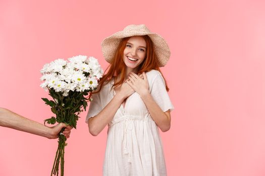 Touched, passionate and lovely alluring redhead female in straw hat, dress, touching heart and sighing with admiration and happiness, flattered for gift, hand extending her bouquet flowers.