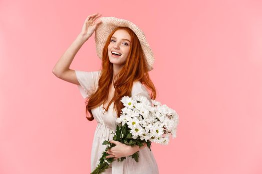 Girl enjoying spring day under sun, stralling buying bouquet for girlfriend, holding white flowers and smiling camera, touching hat cheeky and lovely gazing, standing pink background.
