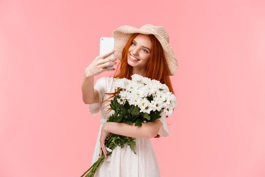 Romantic moments, social media and technology concept. Charming alluring redhead female in dress, straw hat, taking selfie on smartphone applying app filter as holding bouquet of flowers.