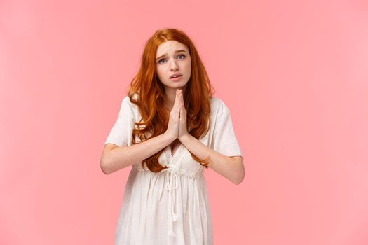 Pleading cute and silly redhead girl acting innocent, press hands together in pray, asking for favour, hopefully looking at camera, frowning gloomy, need something, begging over pink background.