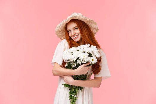 Lovely and alluring romantic cute, silly redhead girl in sun straw hat, embracing beautiful white flowers, receive gift of bouquet and smiling camera cheeky, standing coquettish pink background.