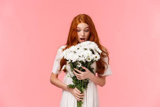 Surprised attractive redhead girl receive romantic valentines day gift, looking at beautiful bouquet of flowers amazed and wondered who might sender be, standing over pink background.